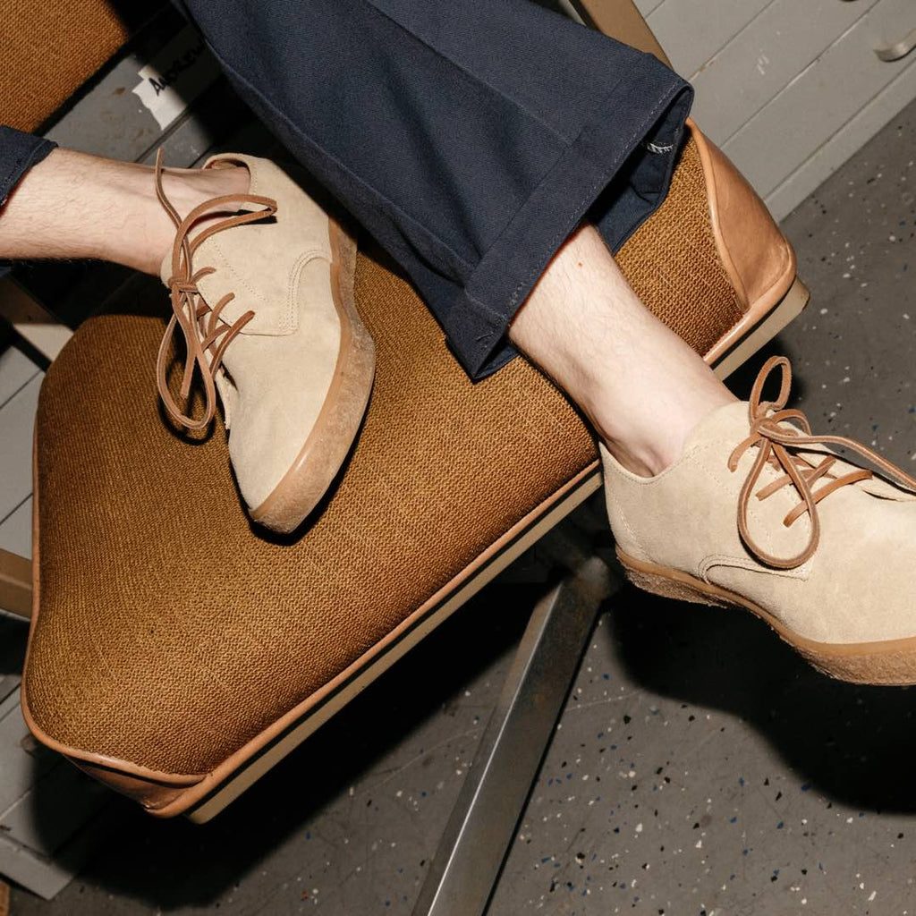 Men's Sun-tans Buck in Sandstone suede, worn with navy pants, feet resting on a tan cushion