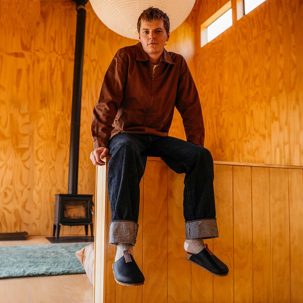 Men's Tiburon Trekker sandal in Black leather, worn by a person seated on a wooden ledge in a cabin