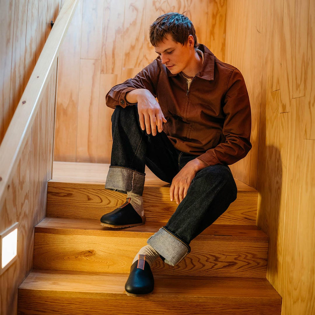Men's Tiburon Trekker sandal in Black leather, worn by a person seated on wooden stairs in a brown jacket