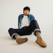 Men's Tiburon Trekker sandal in Maple suede, worn by a person seated on a white floor in a denim jacket