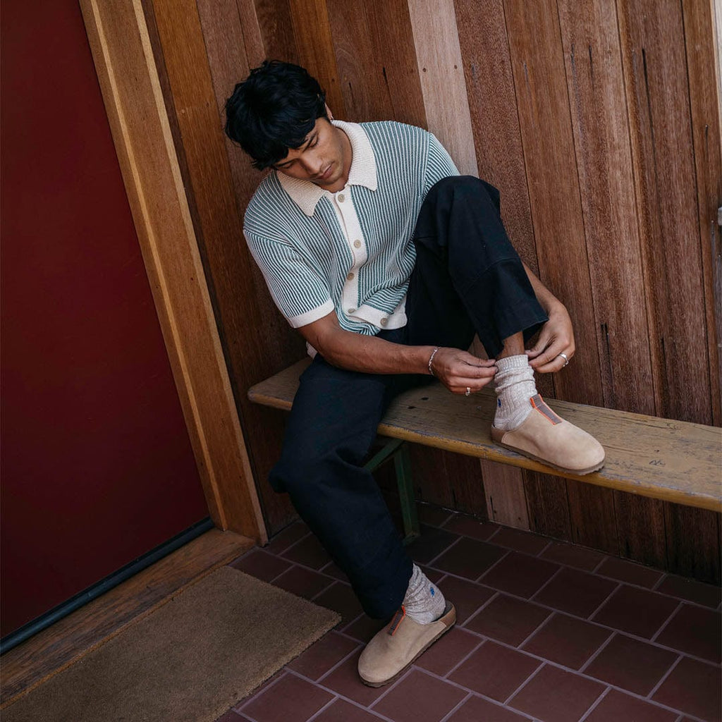 Men's Tiburon Trekker sandal in Maple suede, worn by a person seated on a wooden bench in a small cabin