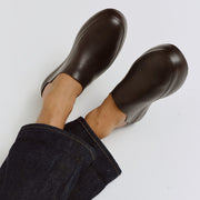 Men's Williams Mule in Dark Chocolate rubber, worn with bare ankles, feet raised, on a white background