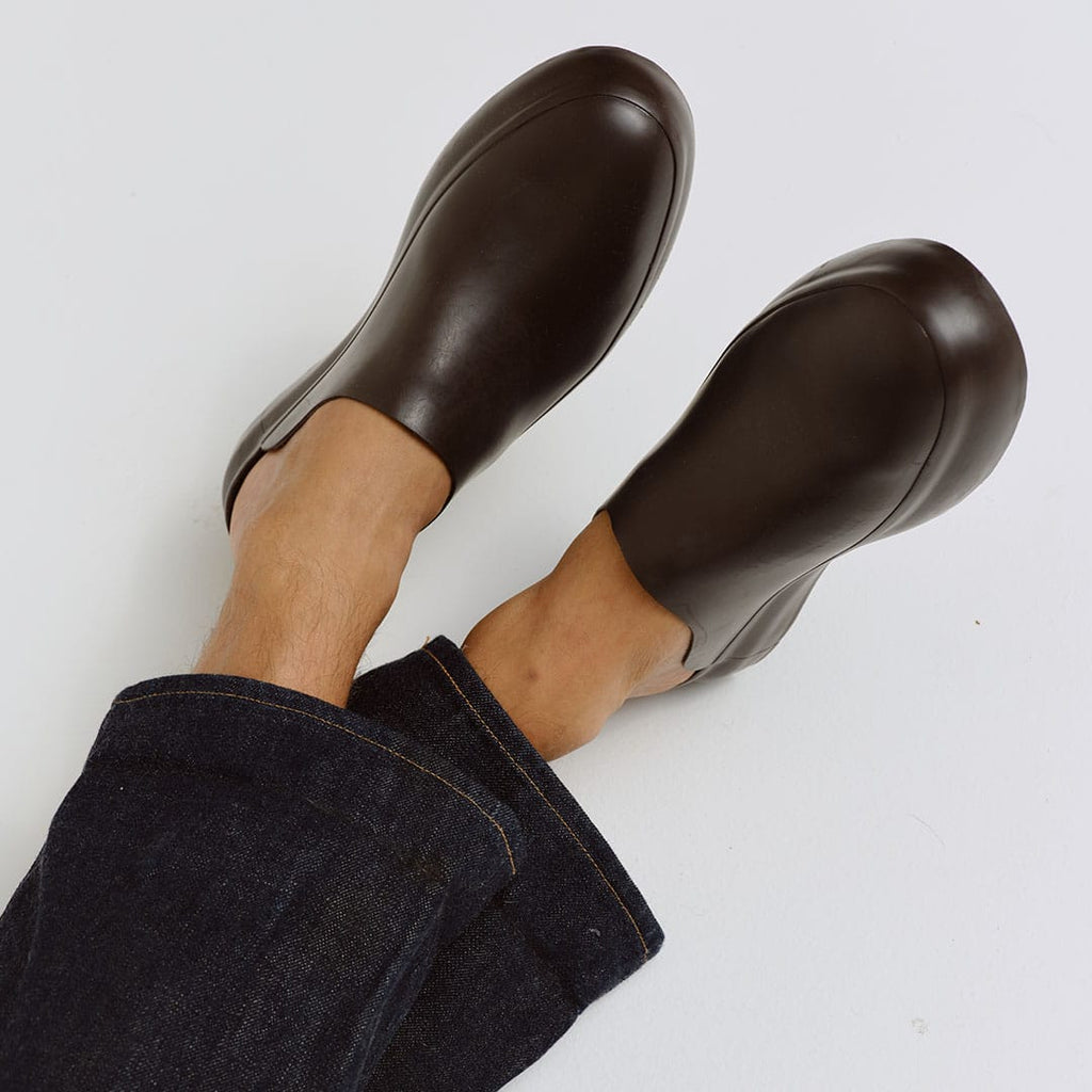Men's Williams Mule in Dark Chocolate rubber, worn with bare ankles, feet raised, on a white background