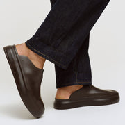 Men's Williams Mule in Dark Chocolate rubber, worn with dark jeans, one foot raised on a white background