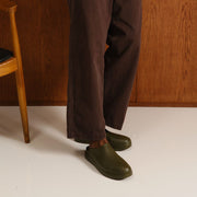 Men's Williams Mule in Utility Olive rubber, worn with brown pants, standing against a wood wall