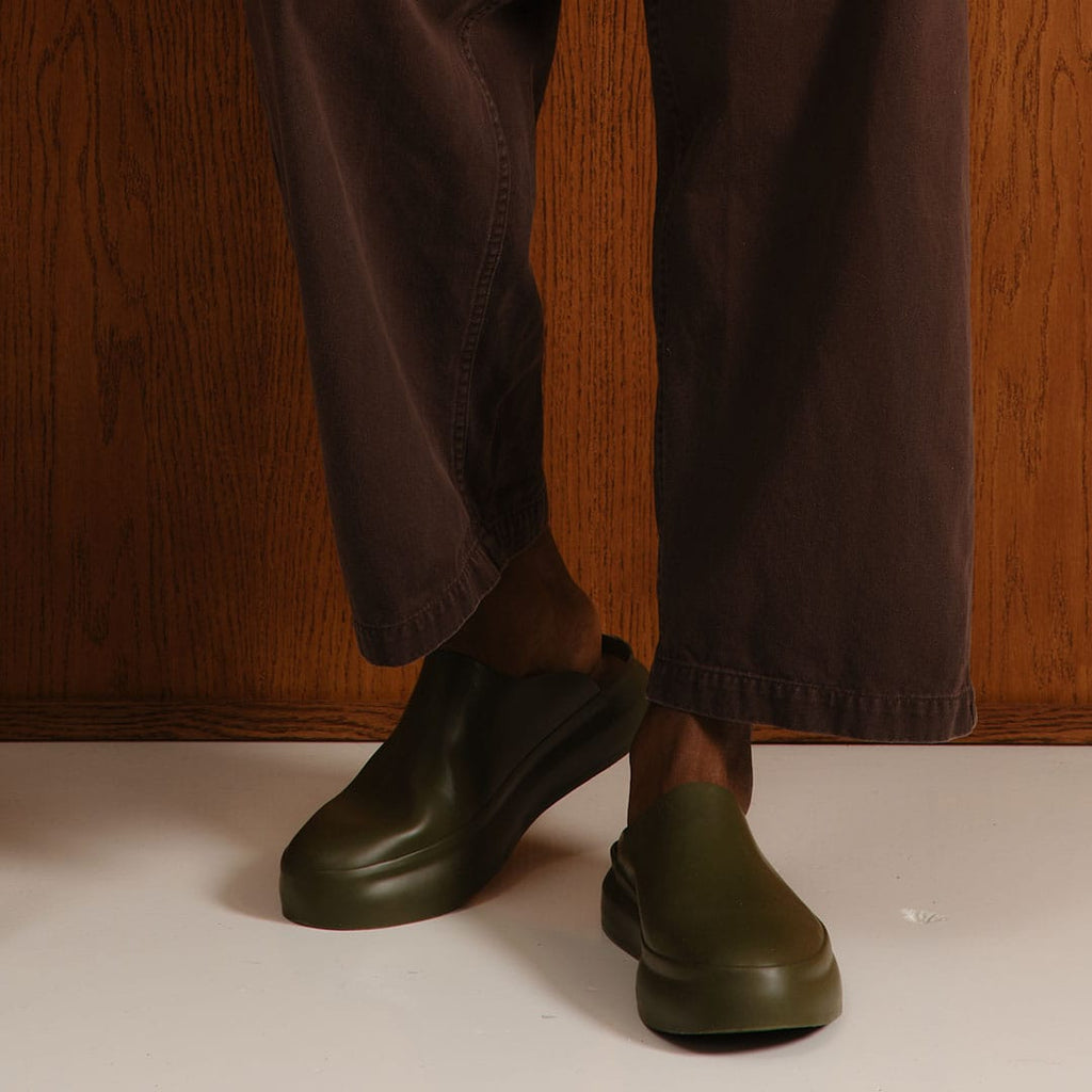 Men's Williams Mule in Utility Olive rubber, worn with dark pants against a wood wall