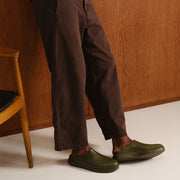 Men's Williams Mule in Utility Olive rubber, worn with brown pants, seated on a wooden chair