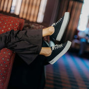 Women's Monterey Sneaker in Black canvas, worn with black pants, feet up on a red patterned couch