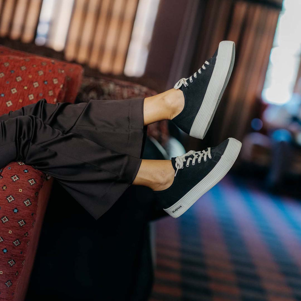 Women's Monterey Sneaker in Black canvas, worn with black pants, feet up on a red patterned couch