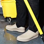 Women's Calistoga Clog in Concrete rubber, worn with black pants in a kitchen beside a mop bucket