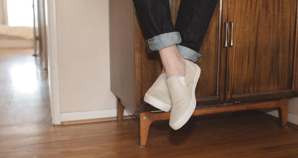 Men's Baja Slip On in Natural canvas, worn with cuffed jeans, standing in a wood-floored hallway