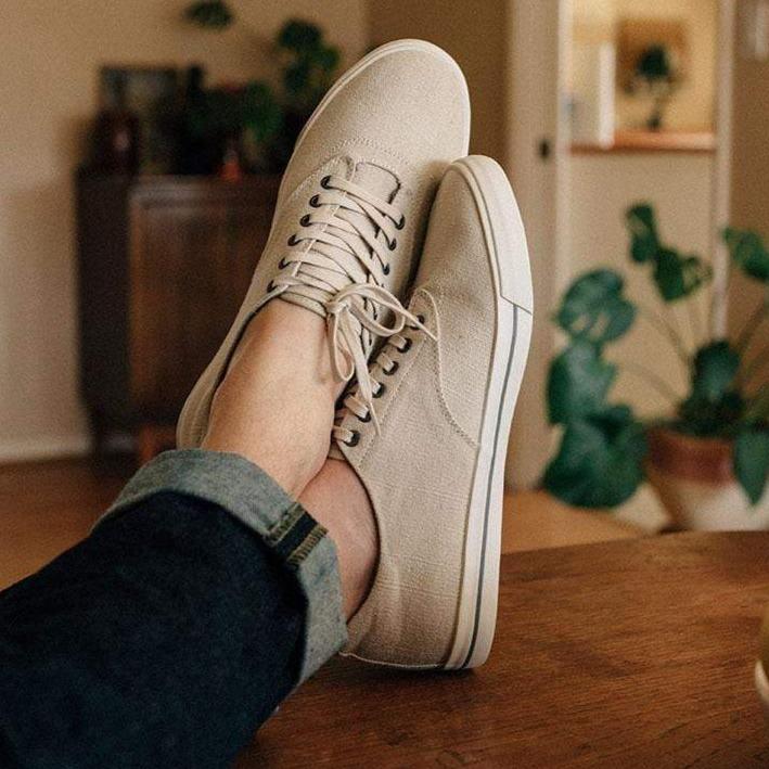 Men's Hermosa Sneaker in Natural canvas, worn with dark cuffed pants, feet resting on a wooden table