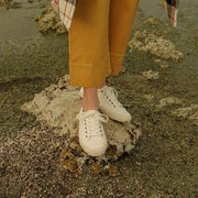 Women's Monterey Sneaker in Bleach canvas, worn with mustard cropped pants, standing on a sandy rock