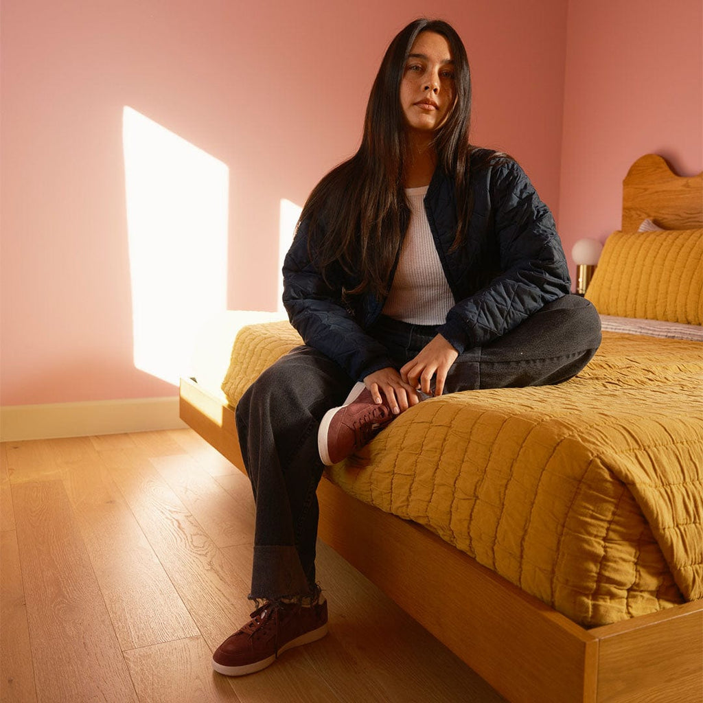 Women's Clipper Sneaker in Plum suede, worn by a woman seated on a bed