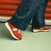 Women's Acorn Trainer sneaker in Candy Apple Red nylon and suede, worn with wide-leg jeans, side view on a teal floor