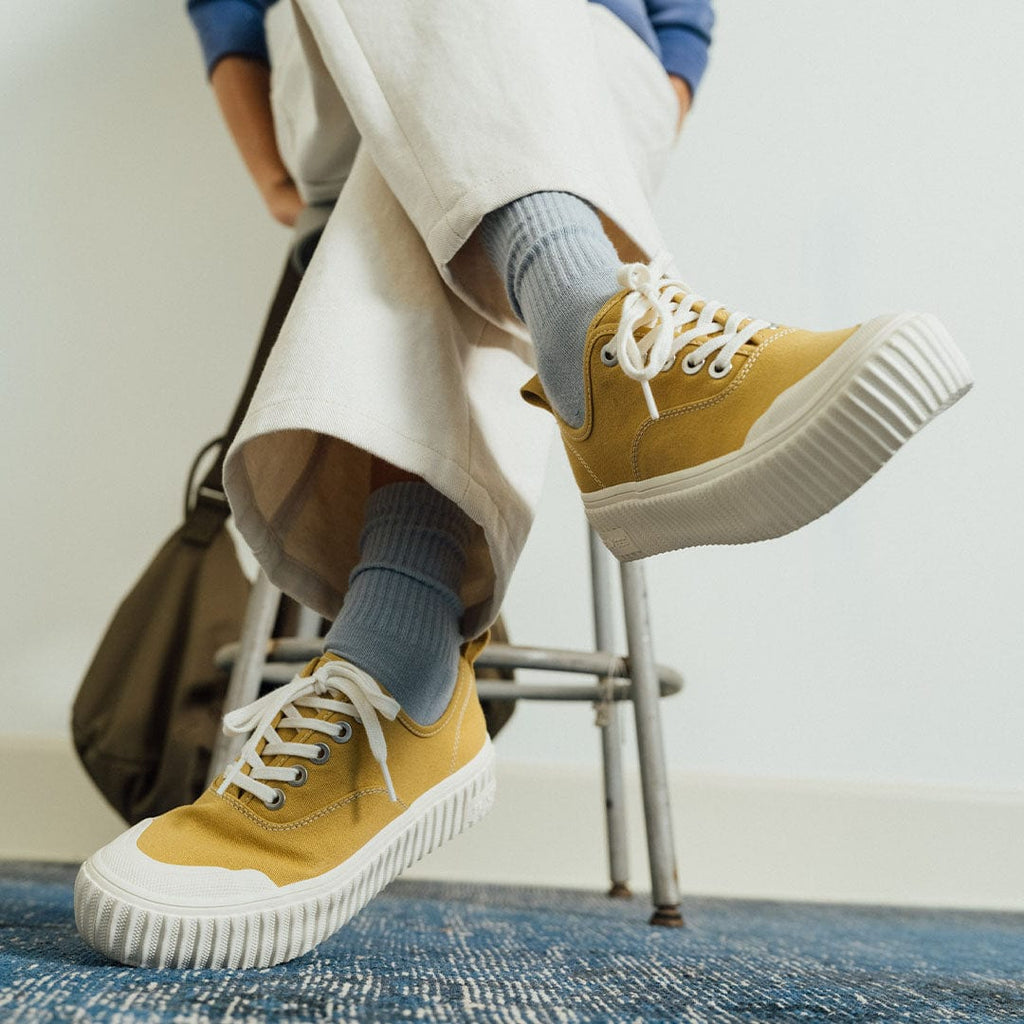 Women's Sato Oxford in Dijon canvas, worn with white pants on a metal stool, side view