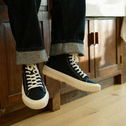 Men's La Brea Lug in Black canvas, worn with cuffed dark jeans, one foot raised, in front of a cabinet