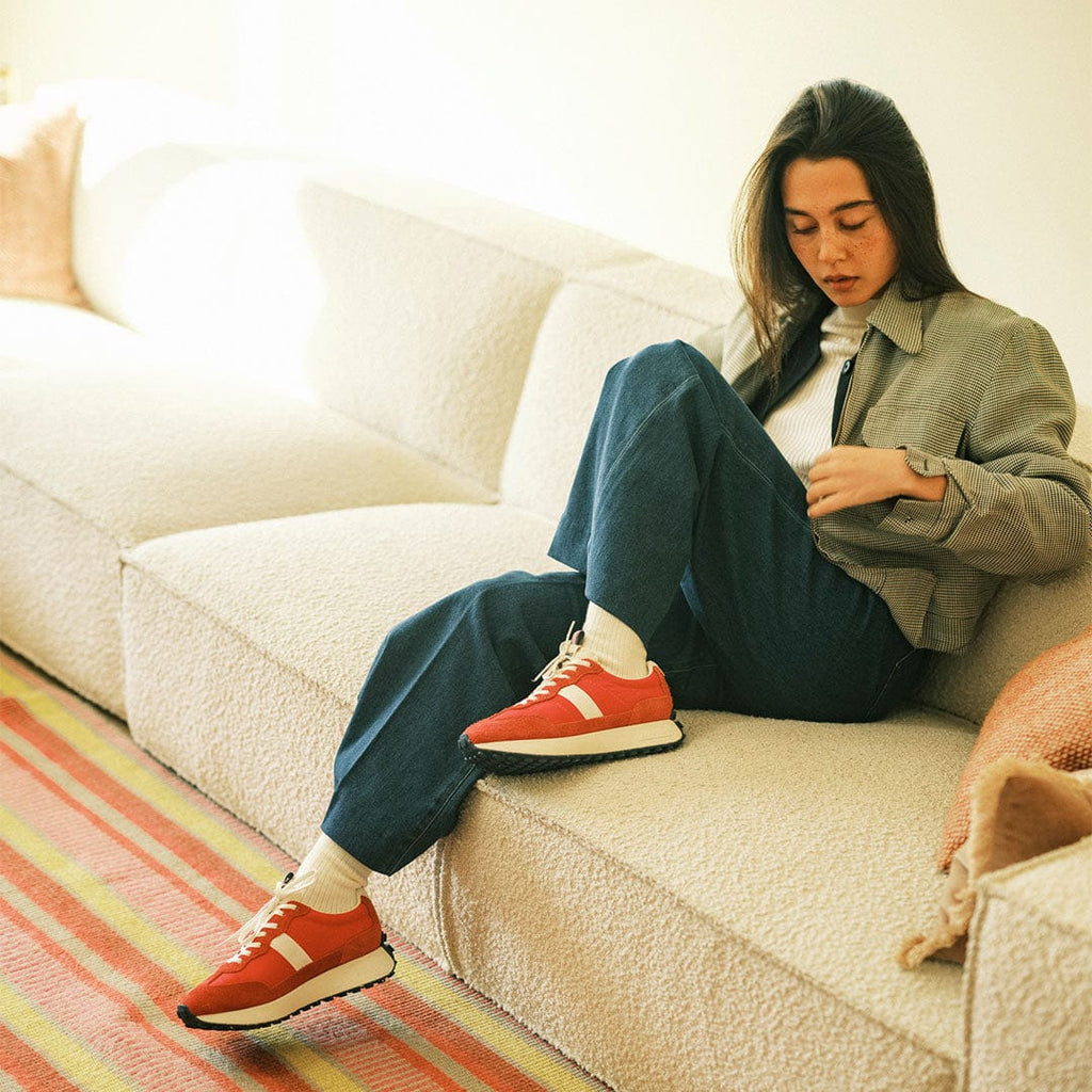 Women's Acorn Trainer sneaker in Candy Apple Red nylon and suede, worn by a woman seated on a cream sofa