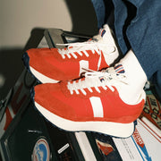 Women's Acorn Trainer sneaker in Candy Apple Red nylon and suede, worn with white socks, stacked on a suitcase