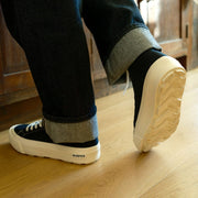Men's La Brea Lug in Black canvas, worn with cuffed dark jeans, one foot raised, showing the cream sole