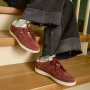 Women's Clipper Sneaker in Plum suede, worn with gray pants on wood stairs, side view