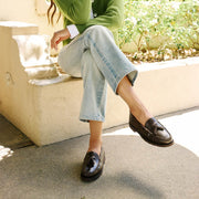 Women's Rickie Tassel Loafer in Dark Chocolate leather, worn with cuffed jeans, seated on a ledge