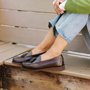 Women's Rickie Tassel Loafer in Dark Chocolate leather, worn with cuffed jeans, feet resting on a wooden bench