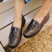 Women's Rickie Tassel Loafer in Dark Chocolate leather, worn with bare feet, seated on a wooden bench