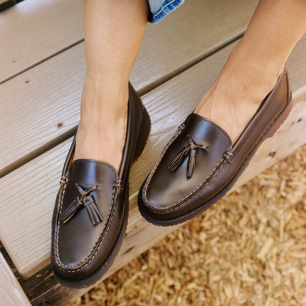 Women's Rickie Tassel Loafer in Dark Chocolate leather, worn with bare feet, seated on a wooden bench