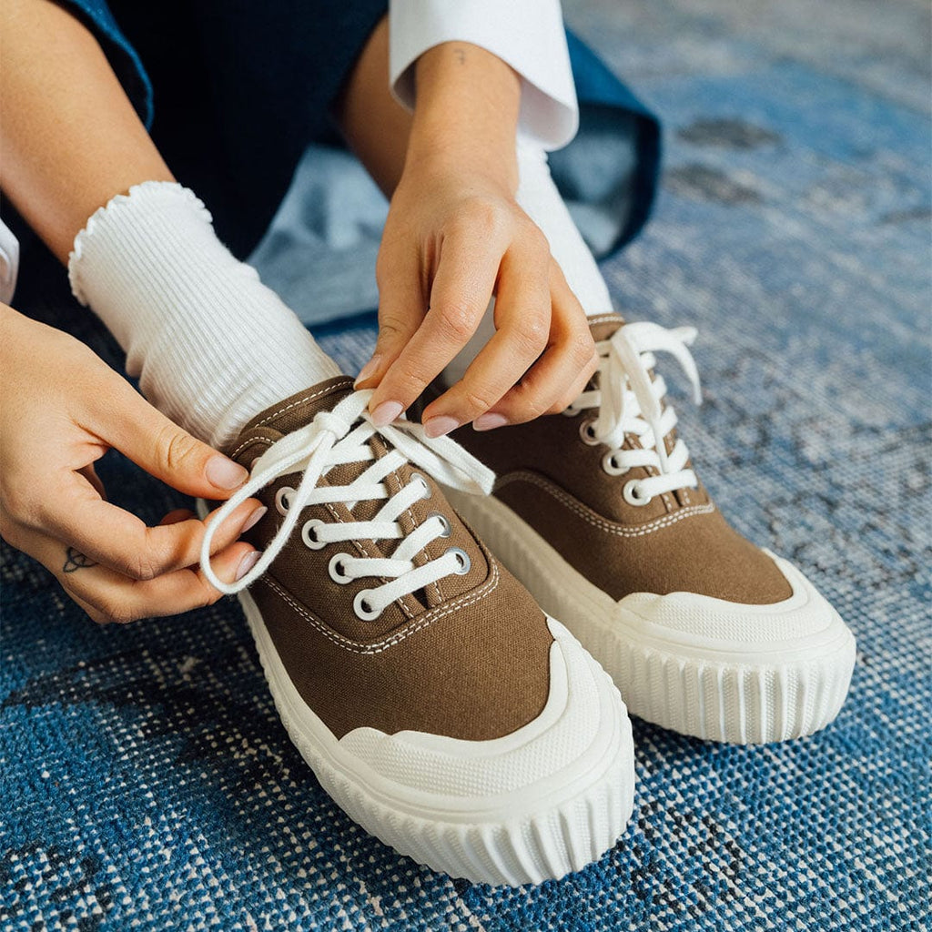 Women's Sato Oxford in Brownstone canvas, close-up of hands tying the white laces on a blue rug
