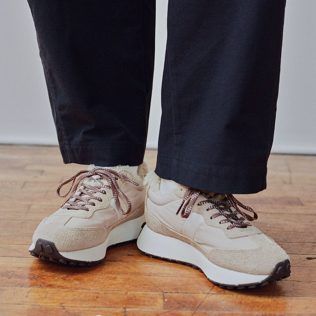 Women's Acorn Trainer sneaker in Cream Cafe nylon and suede, worn with dark pants on a wood floor