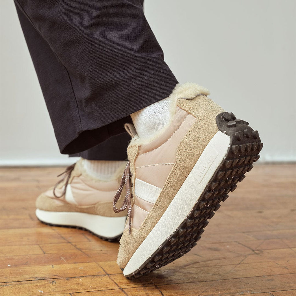 Women's Acorn Trainer sneaker in Cream Cafe nylon and suede, worn with dark pants, one foot raised on a wood floor