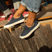 Women's Hawthorne Slip On in True Navy fabric, worn with cuffed jeans, feet resting on a weathered wood fence