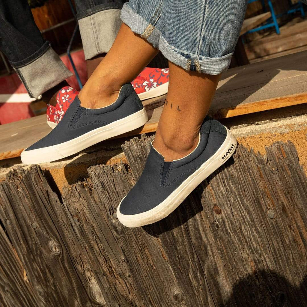 Women's Hawthorne Slip On in True Navy fabric, worn with cuffed jeans, feet resting on a weathered wood fence