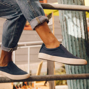Women's Hawthorne Slip On in True Navy fabric, close-up worn with cuffed jeans, foot resting on a rail