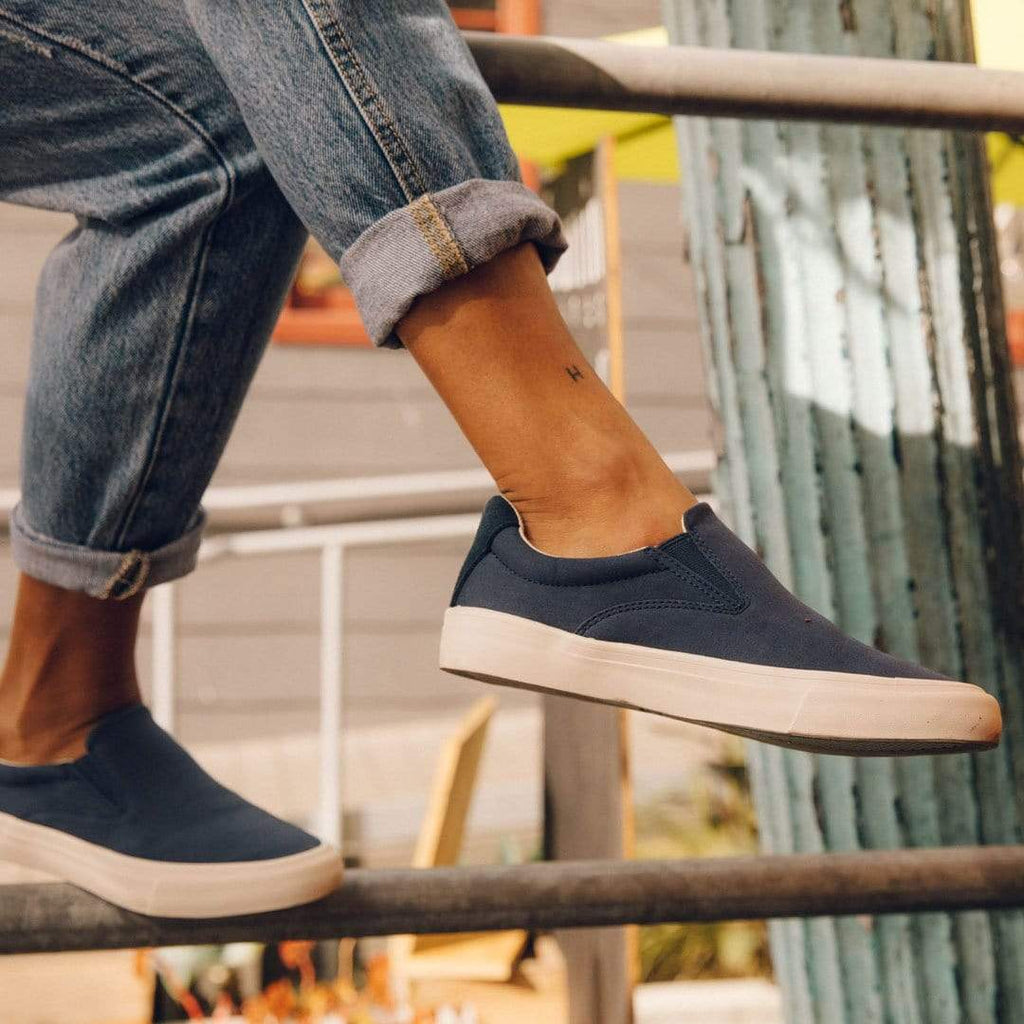 Women's Hawthorne Slip On in True Navy fabric, close-up worn with cuffed jeans, foot resting on a rail
