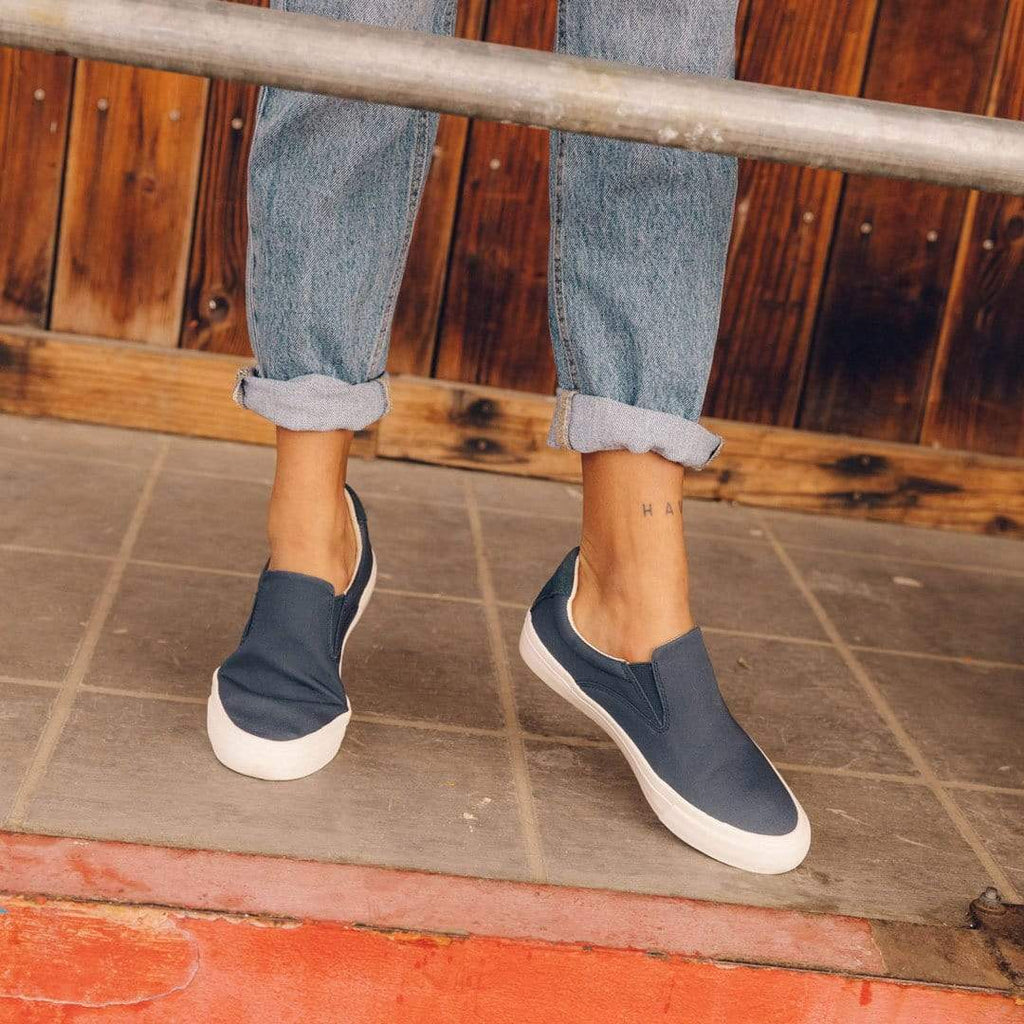 Women's Hawthorne Slip On in True Navy fabric, worn with cuffed jeans, standing on a tiled step