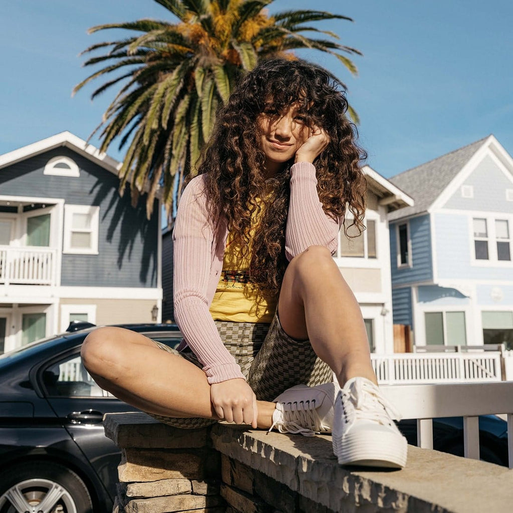 Women's Monterey Sneaker in Bleach canvas, worn with shorts by a person seated on a seawall