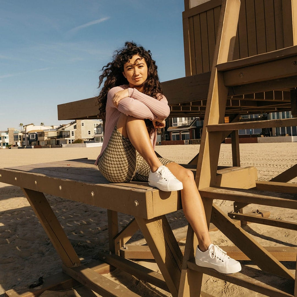 Women's Monterey Sneaker in Bleach canvas, worn with shorts by a person seated at a wooden picnic table on the beach