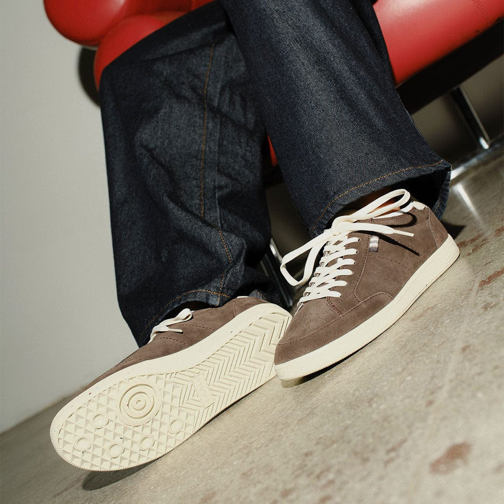 Women's Clipper Sneaker in Espresso suede, worn with dark jeans, one foot on a red chair, side view