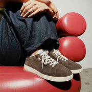 Women's Clipper Sneaker in Espresso suede, worn with dark jeans, foot resting on a red chair, close up