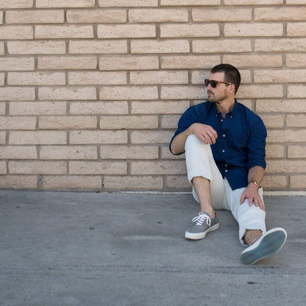 Men's Legend Sneaker in Granite Grey canvas, worn with white pants by a person seated against a brick wall