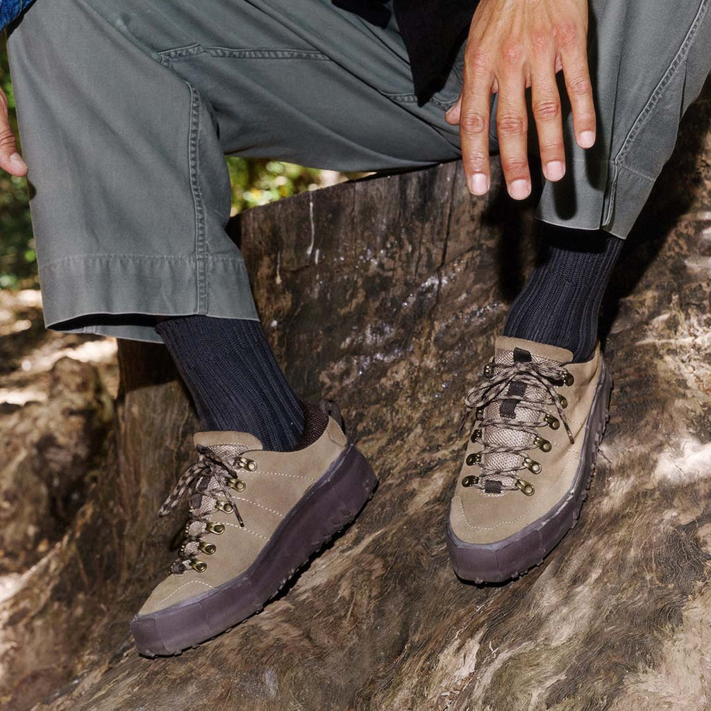 Men's Ridge Urban Hiker hiking sneaker in Desert Palm suede, worn with gray pants and dark socks, seated on a tree stump