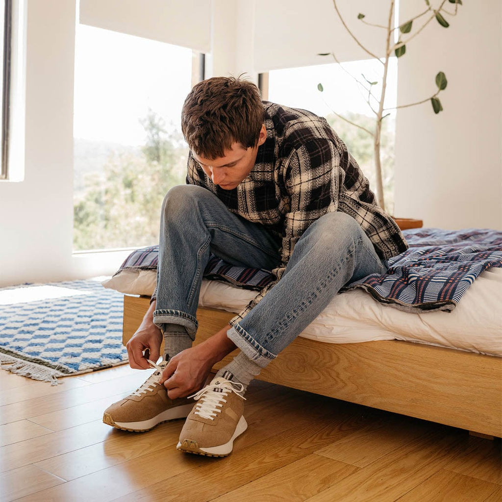 Men's Acorn Trainer sneaker in Tan suede, worn with jeans by a person seated on a bed tying a shoe