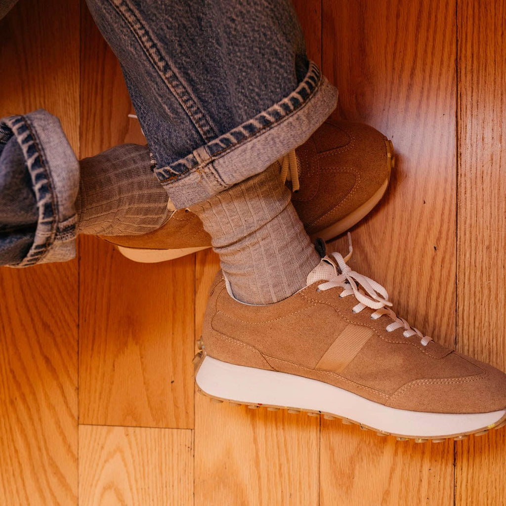 Men's Acorn Trainer sneaker in Tan suede, close-up worn with gray socks on a wood floor