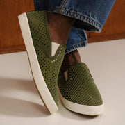 Men's Baja Slip On in Surplus Green vegan nubuck, worn with light jeans, one foot raised, showing the perforated upper