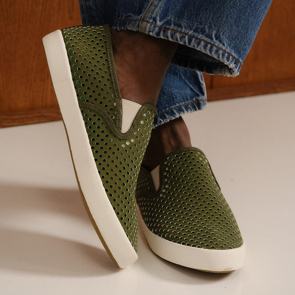 Men's Baja Slip On in Surplus Green vegan nubuck, worn with light jeans, one foot raised, showing the perforated upper