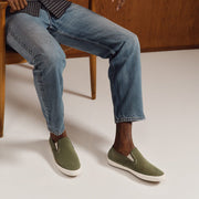 Men's Baja Slip On in Surplus Green vegan nubuck, worn by a person seated in a wooden chair in light jeans