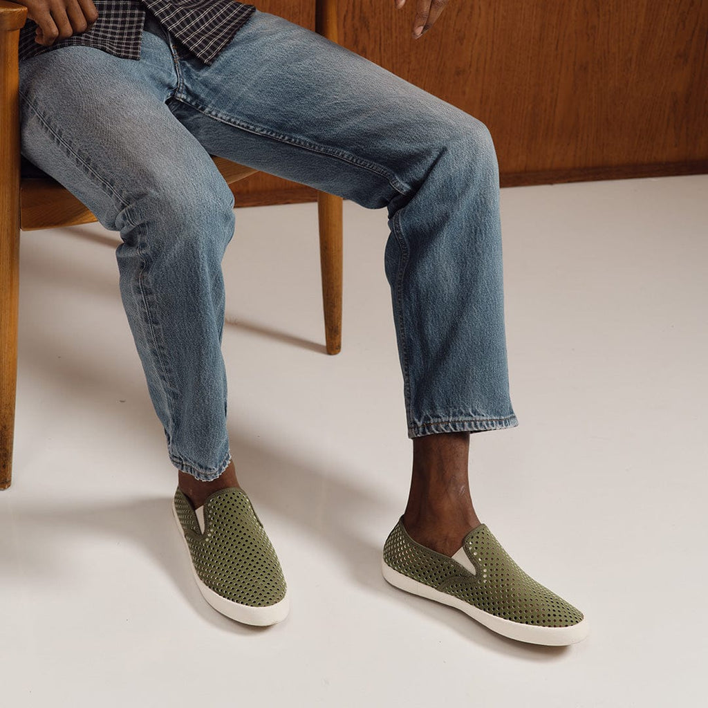 Men's Baja Slip On in Surplus Green vegan nubuck, worn by a person seated in a wooden chair in light jeans