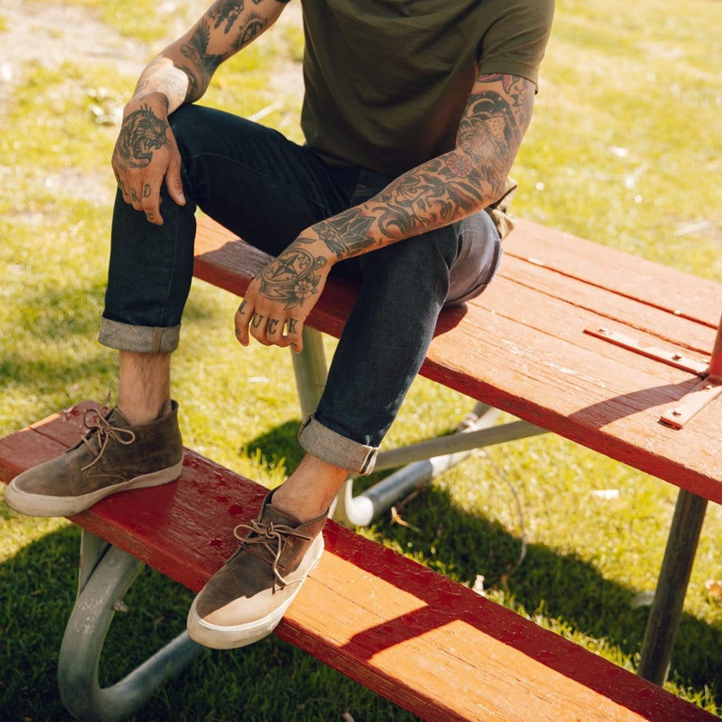 Men's Maslon Desert Boot in Elmwood leather, worn with cuffed jeans by a person seated on a red picnic table
